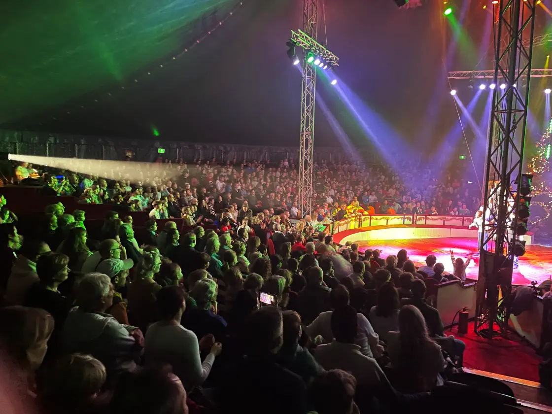 Een volle tribune kijkt naar de piste terwijl groene en paarse spots door de tent zwaaien